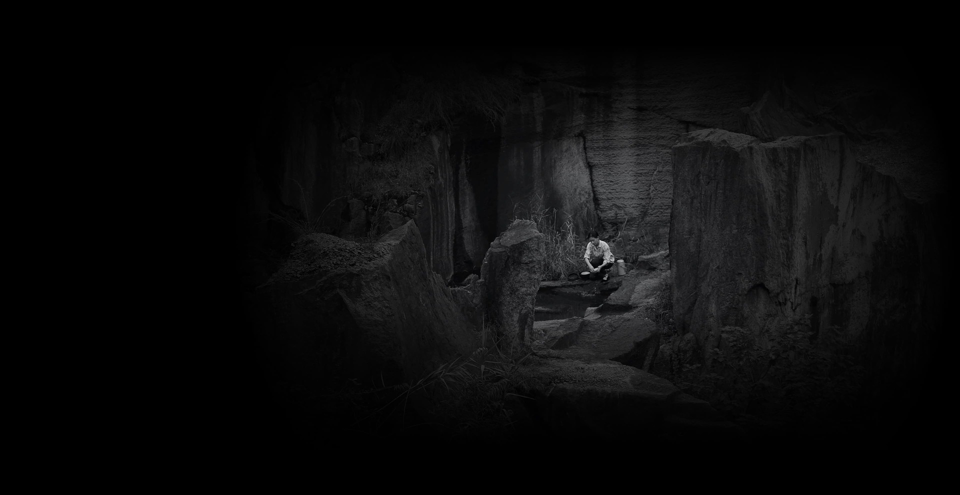 Yang Fudong Introduction background image, a black and white image depicting a woman sitting utop rocks in a craggy environment 