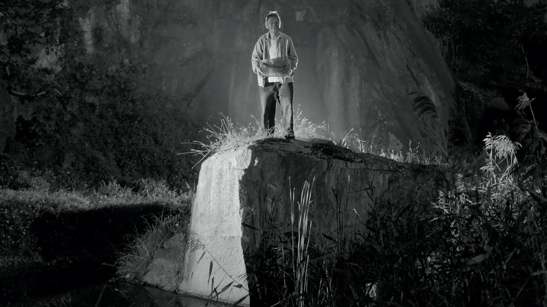 A still from 'Moving Mountains' - Depicting an old man standing atop a ledge in a cave, black and white