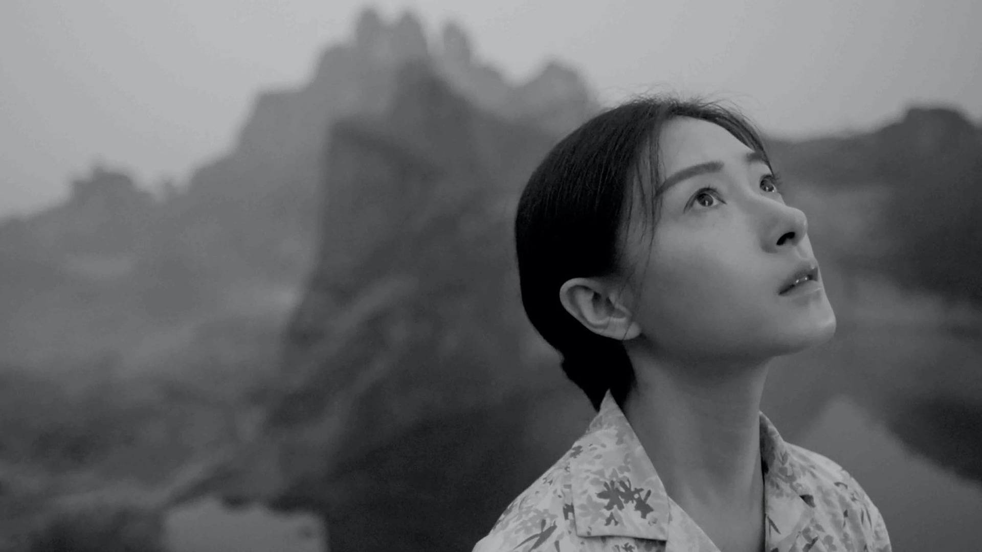 A still from 'Moving Mountains' -  depicting a woman looking forlornly towards the sky, with mountains in the background. Image is in grayscale..