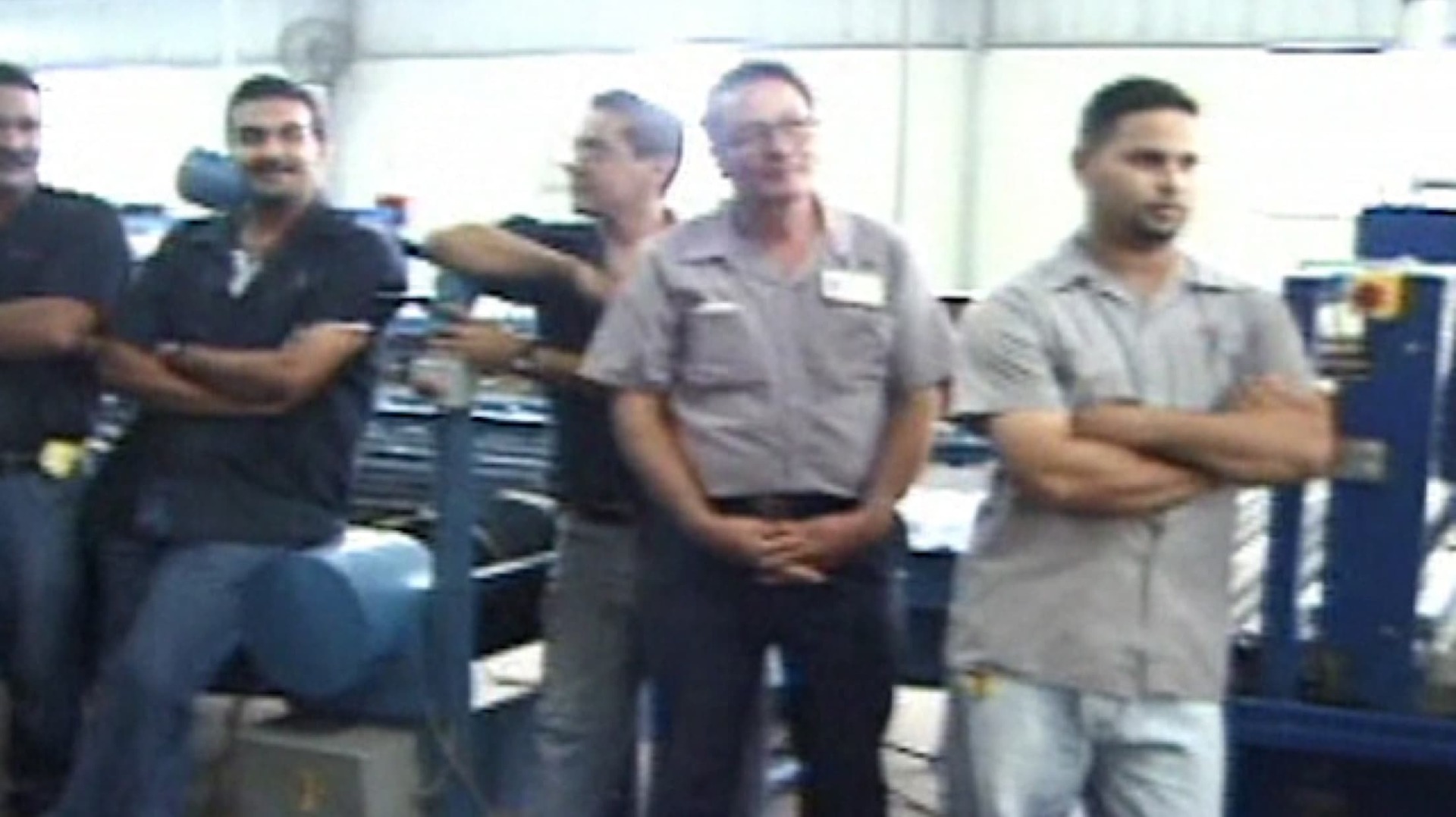 Still from 'Fabrica' film - shows male employees standing in a meeting in a factory