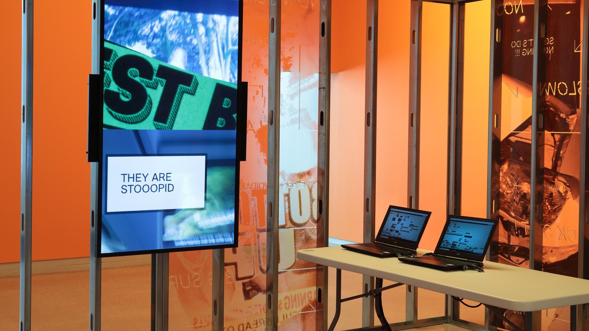 Interior shot of 'Shame Space' shows a screen containing a typographic piece of artwork with two laptops