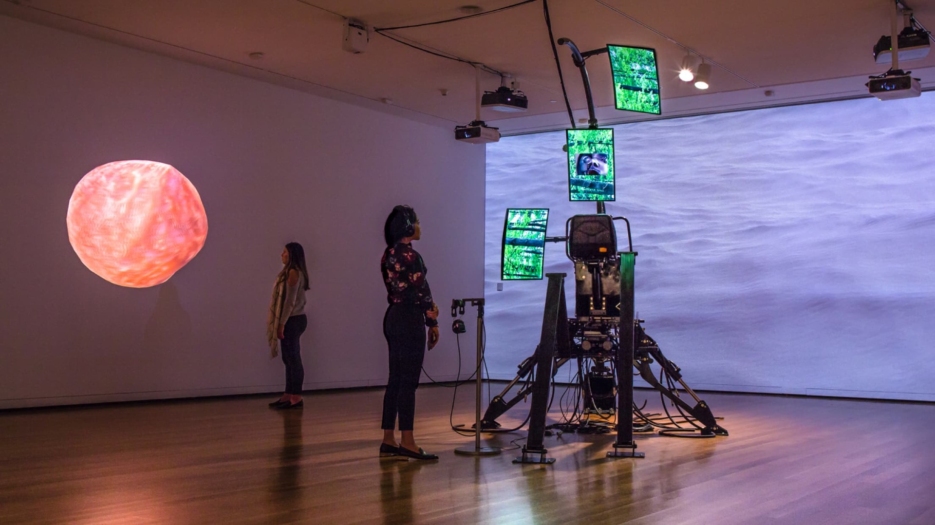 A still from 'Inhabitality' - depicts a woman listening to a soundscape matched with a set of three monitors in a rig of computers, with two projector backgrounds