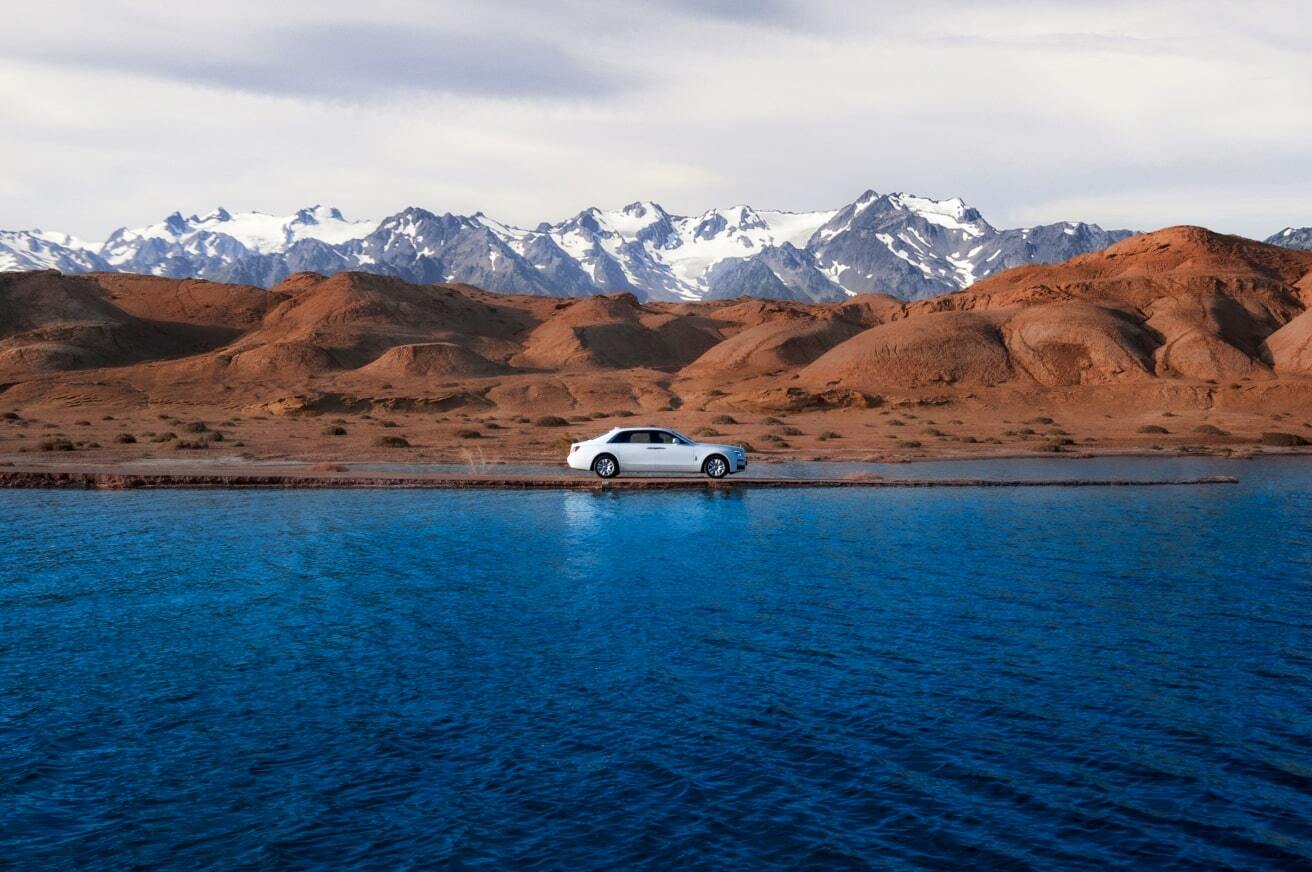 A shot of a beautiful landscape with a lake, mountains and a Rolls-Royce Phantom parked in the middle