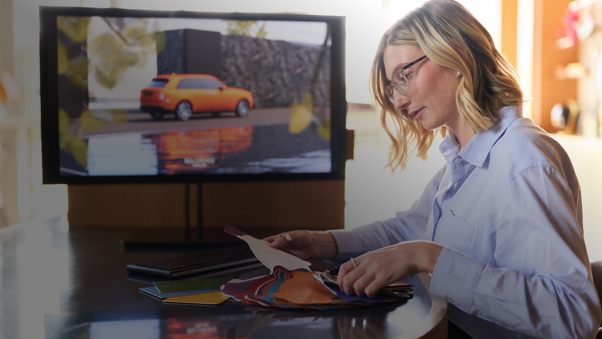 A woman sitting at a table looking at a color swatch with an orange Rolls-Royce Cullinan displayed in a screen in the back