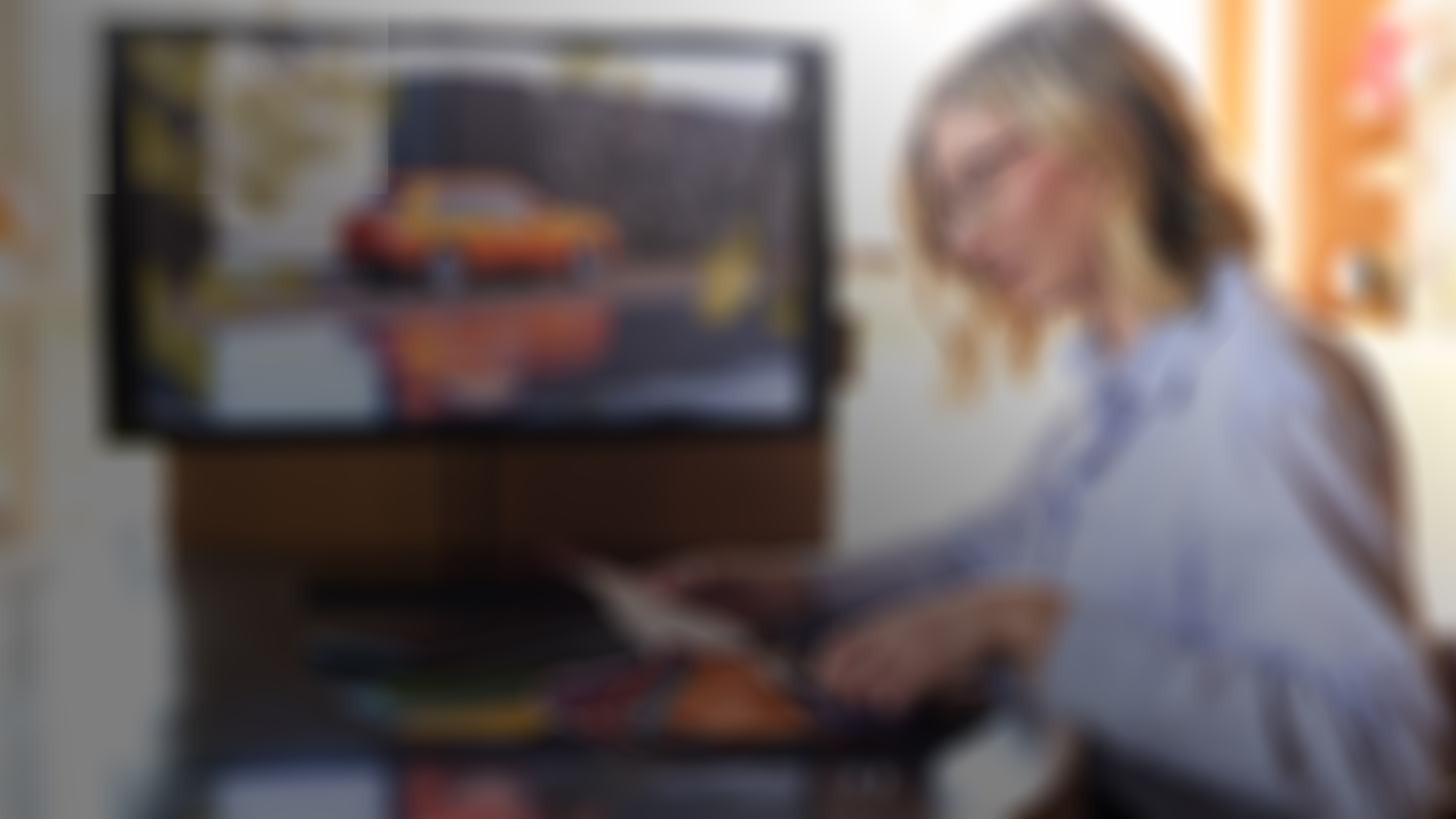 A woman sitting at a table looking at a color swatch with an orange Rolls-Royce Cullinan displayed in a screen in the back