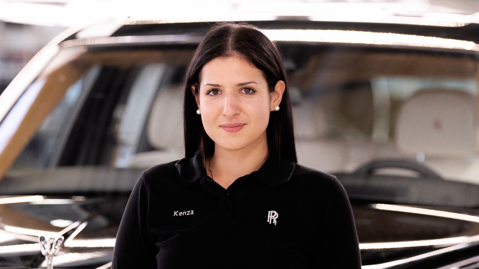 A young Rolls-Royce graduate wearing a polo shirt with the name Kenza and the Rolls-Royce monogram on it and a Rolls-Royce car in the background