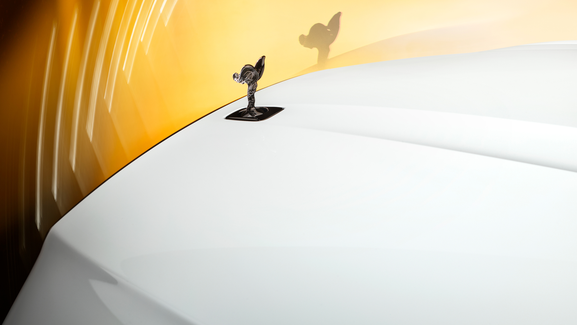 Angled shot of the Spirit of Ecstasy on a white Rolls Royce Black Badge, with yellow lighting backdrop.