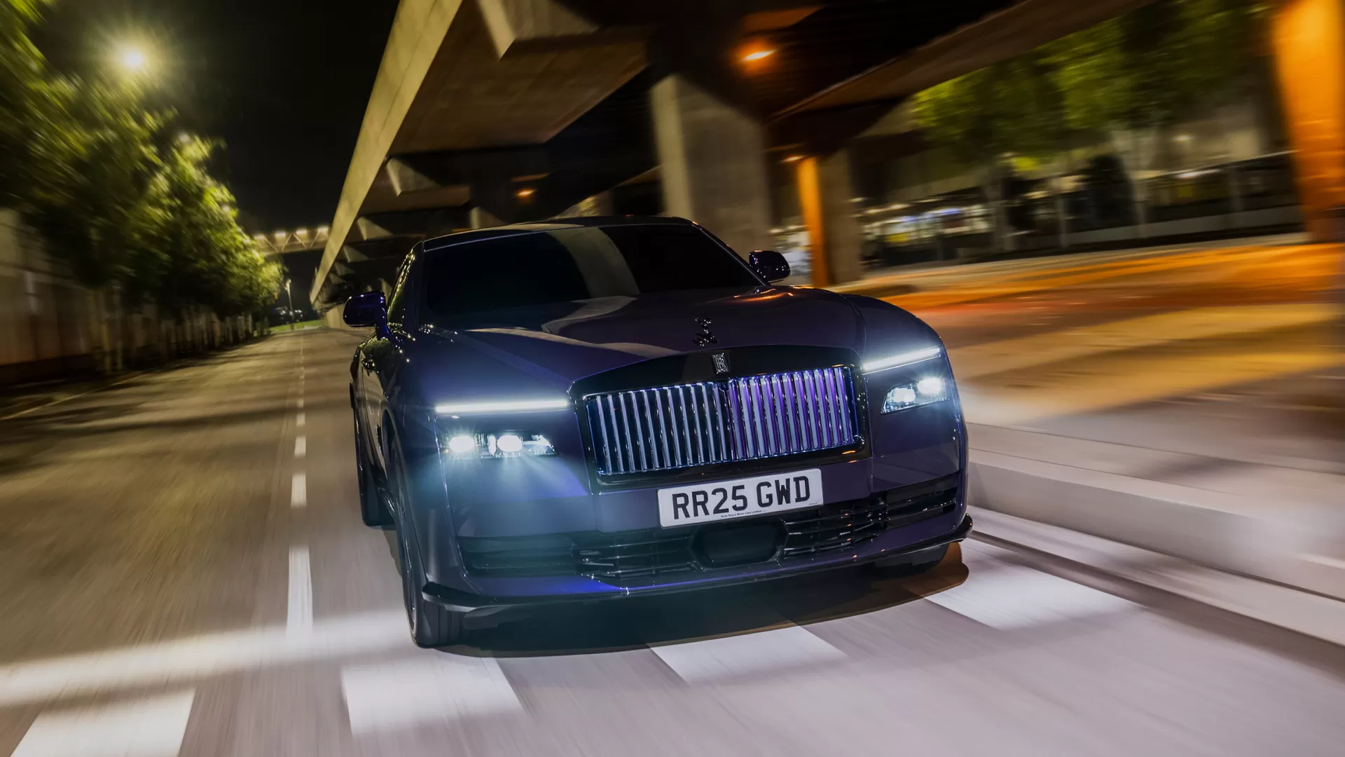 Purple Black Badge Spectre driving quickly through the street at night. A front side angled shot showcasing the classic headlights. 