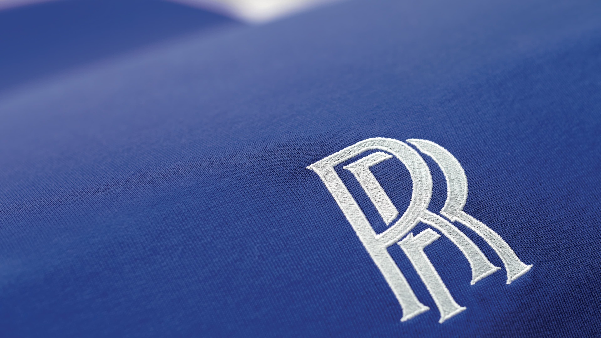 A close-up shot of a white Rolls-Royce Motor Car monogram stitched into the tailored indoor cover in blue
