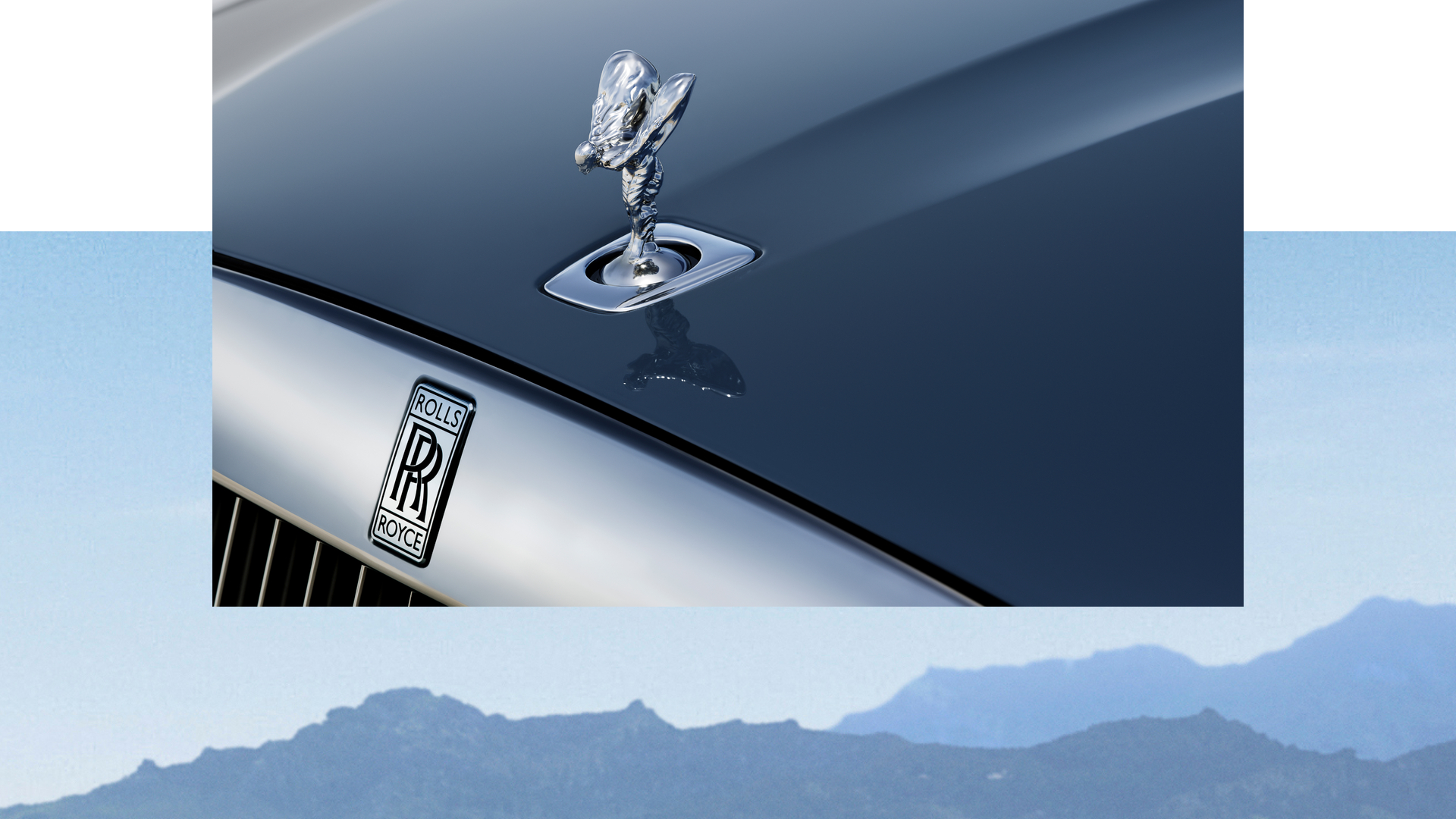 Close up of a silver spirit of ecstasy on a navy blue Ghost Extended SII. A blue and white background drop behind it of mountains. 