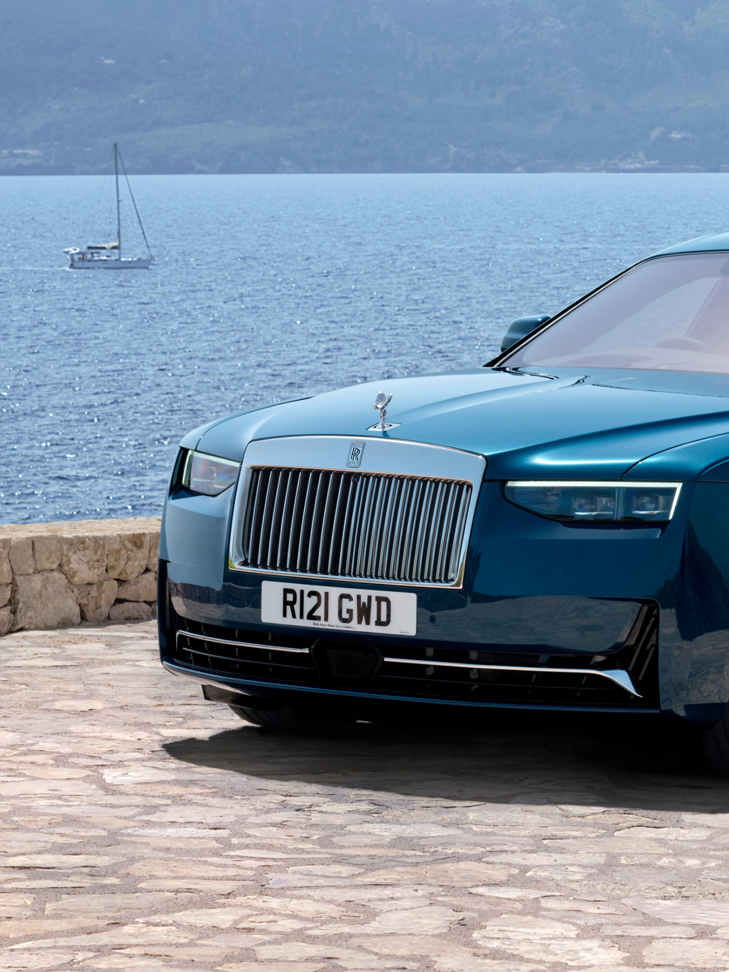 Image of the front half of the car including the grill, sitting beside the ocean with a sailboat in the background drifting away.